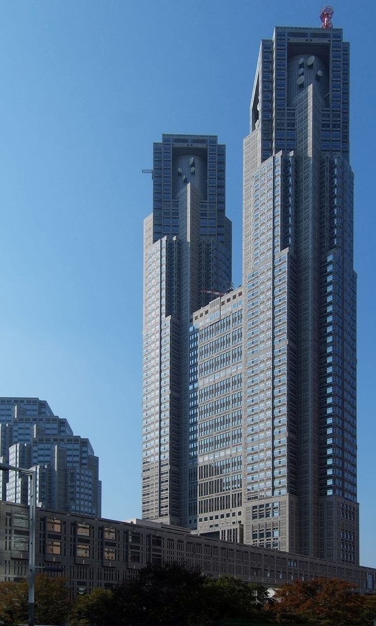 Комплекс зданий мэрии в ТокиоКомплекс зданий Токийской мэрии является визитной карточкой столицы Японии. Он спроектирован известным японским архитектором Кендзо Танге, одним из основоположников направления «метаболизма» в архитектуре. Ансамбль наглядно демонстрирует симбиоз модернизма и традиционного японского стремления к целостности и созерцательности. На воплощение в жизнь грандиозного проекта ушло более трех лет, строительство было окончено в 1991 году. На его возведение затрачено около 1 миллиарда долларов.Токийский муниципалитет состоит из трёх зданий, объединенных в единую архитектурную композицию: Токийское правительственное здание №1, Токийское правительственное здание №2 и Дом народного собрания. Высота первого, главного, здания почти 243 метра (45 этажей), также оно включает три подземных уровня. Второе — немного ниже (34 этажа и три поземных). Третье имеет всего восемь этажей, один из которых находится ниже уровня земли; оно расположено чуть поодаль от двух основных, напротив первого корпуса. Все три здания объединены между собой узкими «мостами». В центре образовавшегося квадрата расположена площадь в форме веера, вокруг разбит сквер.Главное здание напоминает футуристический готический собор — на высоте 33-го этажа оно раскалывается на две башни — Северную и Южную. Эти башни неофициально окрестили "японским Нотр-Дам де Пари". В наружном и внутреннем дизайне Токийского муниципалитета используются элементы, напоминающие компьютерные микросхемы. В частности, изображения микропроцессоров находятся на потолке второго этажа главного корпуса, а также в других частях здания.Здания возведены по технологиям сейсмостойкого строительства. Конструкция способна выдерживать 8-балльное землетрясение. Воздействие силы ветра удалось сократить на треть путём правильной ориентации здания в пространстве. Использование округлых форм с наветренной стороны и грубой текстуры с подветренной, расположение наклона крыш высотных башен под углом 45 градусов и наличие высоких деревьев в окрестности, позволило снизить ветровые нагрузки.